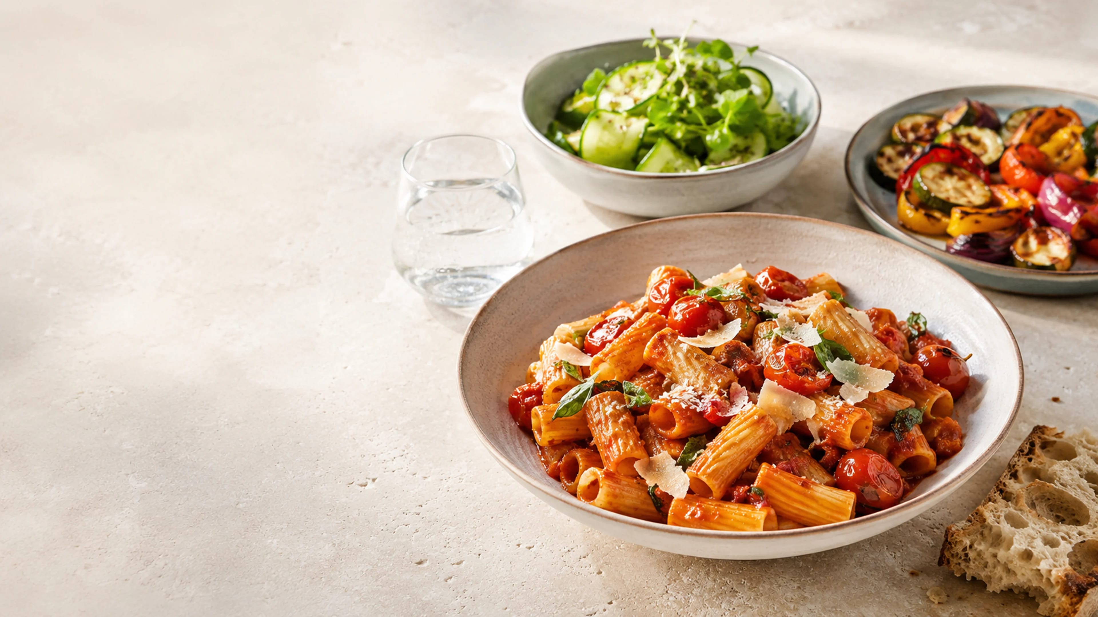 Rigatoni mit Tomatensauce, Salat und Ofengemüse auf einem hellen Tisch.