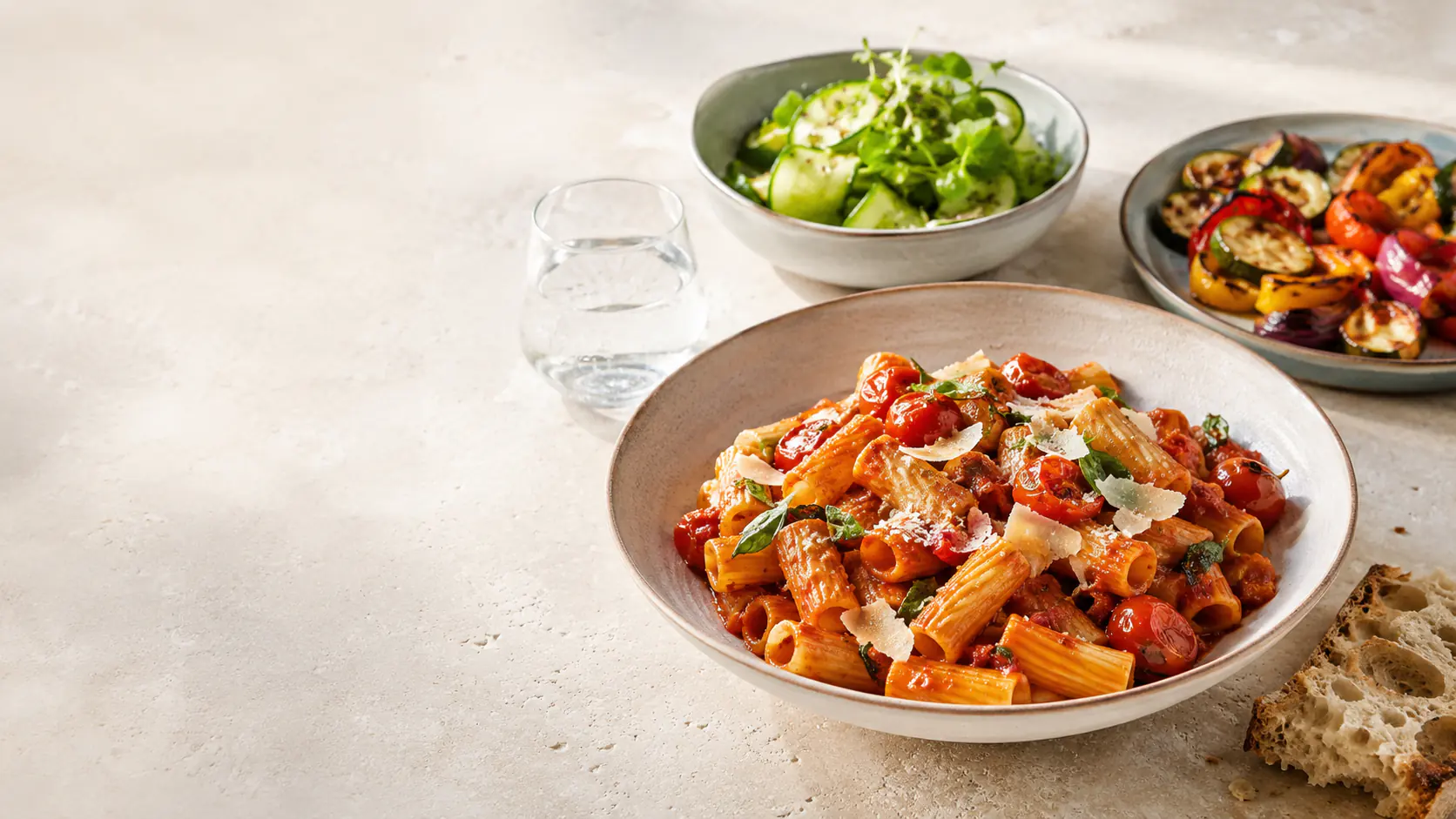 Rigatoni mit Tomatensauce, Salat und Ofengemüse auf einem hellen Tisch.